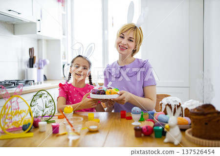 A mother and daughter wearing bunny ears happily decorate colorful Easter eggs together in a bright kitchen setting 137526454