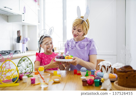 A mother and daughter wearing bunny ears are happily painting and decorating Easter eggs together at a kitchen table 137526455
