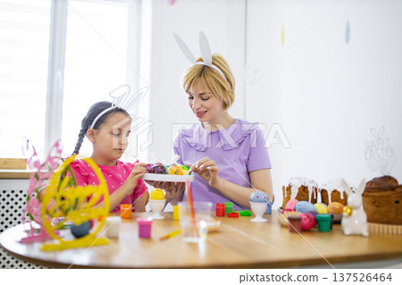 A mother and her daughter wearing bunny ears are happily decorating colorful Easter eggs together at a wooden table filled with festive treats and craft supplies A mother and her daughter wearing bunny ears are happily decorating colorful Easter eggs together at a wooden table filled with festive treats and craft supplies 137526464