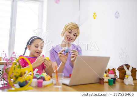 A mother and daughter wearing bunny ears paint Easter eggs together while following an online tutorial on a laptop A mother and daughter wearing bunny ears paint Easter eggs together while following an online tutorial on a laptop 137526466