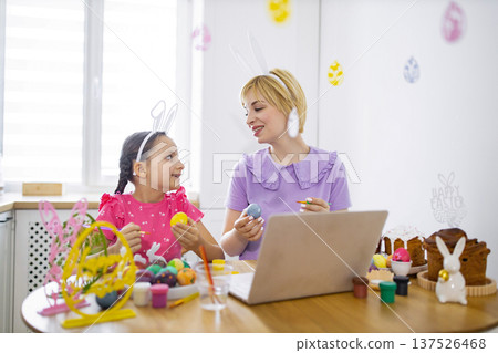 A mother and daughter wearing bunny ears are painting Easter eggs together at a table with a laptop and Easter cakes A mother and daughter wearing bunny ears are painting Easter eggs together at a table with a laptop and Easter cakes 137526468
