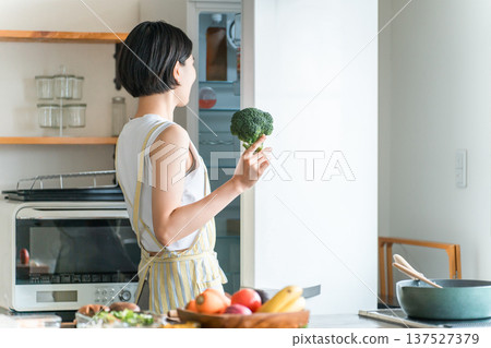 一位年輕的亞洲女性/家庭主婦，留著短髮，繫著圍裙，在廚房裡拿著蔬菜 137527379