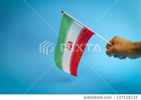 Iran flag held by a human hand isolated on a blue background. Conceptual photography representing Iranian protests, geopolitical crisis, and the internal struggle for freedom and human rights 137528135