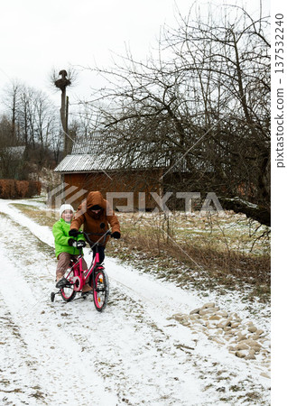 Joyful winter adventure two children ride a pink bicycle through a snow-covered countryside path surrounded by rustic charm 137532240