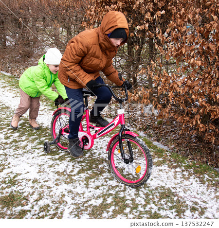 Children enjoy a winter day, biking through a snowy park while one friend gives a helping push under the muted sky of early afternoon Children enjoy a winter day, biking through a snowy park while one friend gives a helping push under the muted sky of early afternoon 137532247