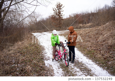 Two children enjoying a winter day on a snowy path while one inspects a pink bicycle amidst a serene nature backdrop in a quiet outdoor location Two children enjoying a winter day on a snowy path while one inspects a pink bicycle amidst a serene nature backdrop in a quiet outdoor location 137532249