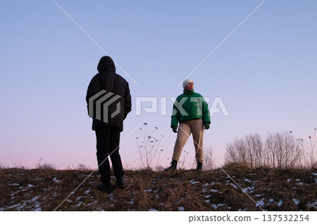 Two figures on a winter evening hillside embracing the cool breeze while the sky transitions to twilight in a serene landscape Two figures on a winter evening hillside embracing the cool breeze while the sky transitions to twilight in a serene landscape 137532254
