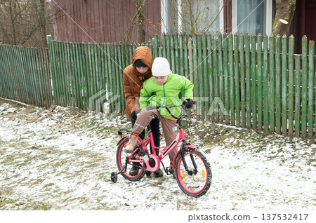 Two children enjoying a winter afternoon as one teaches the other how to ride a pink bicycle on a snowy lawn beside a rustic wooden fence Two children enjoying a winter afternoon as one teaches the other how to ride a pink bicycle on a snowy lawn beside a rustic wooden fence 137532417