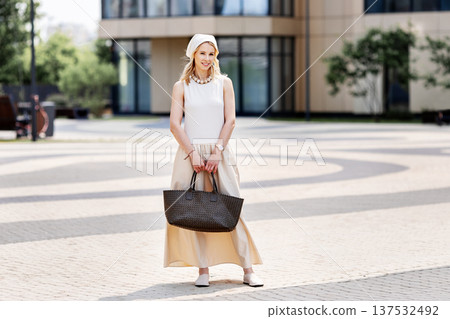 Full length portrait stylish young woman standing outdoors in city near modern building. Female wearing beige sleeveless maxi dress, headscarf, flat shoes, holding Tote handbag, summer urban fashion Full length portrait stylish young woman standing outdoors in city near modern building. Female wearing beige sleeveless maxi dress, headscarf, flat shoes, holding Tote handbag, summer urban fashion 137532492