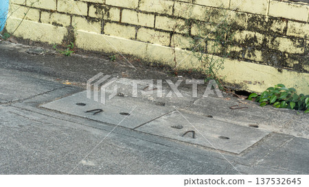 High angle view of concrete drainage manhole covers on a city street in Thailand. Urban infrastructure and sewage system along a weathered yellow brick wall in a residential area. High angle view of concrete drainage manhole covers on a city street in Thailand. Urban infrastructure and sewage system along a weathered yellow brick wall in a residential area. 137532645