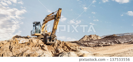 An excavator moves dirt at a construction site in a dry, rocky area during the afternoon. The sky is clear and the ground is uneven An excavator moves dirt at a construction site in a dry, rocky area during the afternoon. The sky is clear and the ground is uneven 137533556