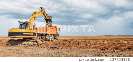 A yellow excavator loads dirt into a large orange truck on a construction site. The site is wide open with dark clouds forming in the sky 137533558