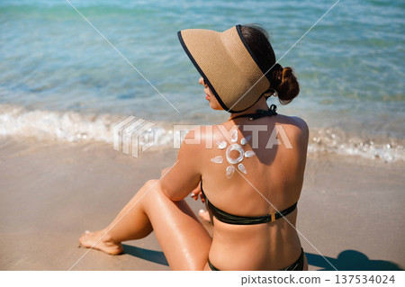 Beautiful woman relaxing on beach with suncream sun pattern on her back holding sunscreen lotion during summer vacation Beautiful woman relaxing on beach with suncream sun pattern on her back holding sunscreen lotion during summer vacation 137534024