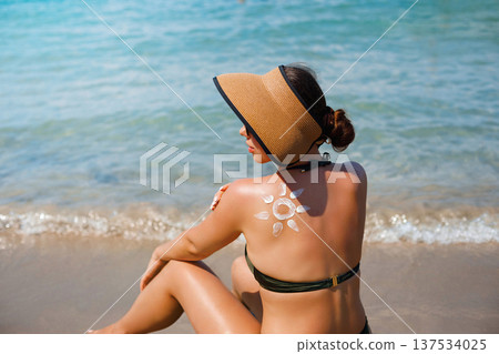 Woman sitting on Mediterranean beach holding sunscreen lotion with decorative suncream sun pattern on her back 137534025