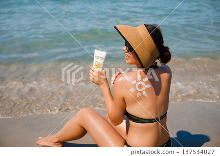 Woman relaxing on sandy beach showing suncream sun pattern on her back while holding sunscreen lotion Woman relaxing on sandy beach showing suncream sun pattern on her back while holding sunscreen lotion 137534027