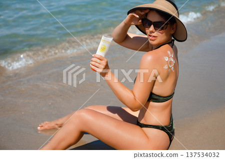 Woman Applying Sun Cream on Tanned Shoulder In Form Of The Sun. Sun Protection.Sun Cream. Skin and Body Care. Girl Using Sunscreen to Skin. Female Holding Suntan Lotion. Woman Applying Sun Cream on Tanned Shoulder In Form Of The Sun. Sun Protection.Sun Cream. Skin and Body Care. Girl Using Sunscreen to Skin. Female Holding Suntan Lotion. 137534032