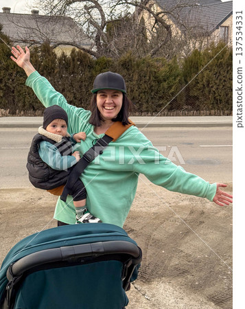 Happy mother with baby carrier and stroller on suburban street Happy mother with baby carrier and stroller on suburban street 137534331