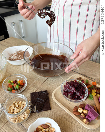 A woman in the kitchen preparing chocolate dough in a glass bowl. Chocolate, candied fruits, and spices on the table A woman in the kitchen preparing chocolate dough in a glass bowl. Chocolate, candied fruits, and spices on the table 137534404