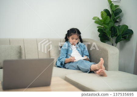 Young girl sitting comfortably on a couch in a cozy living room, engaged in drawing or writing in a notebook with a laptop nearby Young girl sitting comfortably on a couch in a cozy living room, engaged in drawing or writing in a notebook with a laptop nearby 137535695