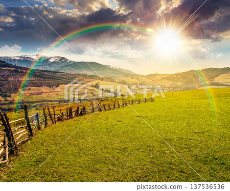 sunset over Springtime rural landscape. Wooden fence along the path through agricultural fields in mountains. beautiful countryside scenery under cloudy sky with rainbow. remote alpine pasture 137536536