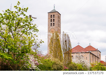 Catholic church Unsere Liebe Frau in Wuerzburg, Germany, showing the stone clock tower framed by green spring trees and blooming branches under a cloudy sky. Catholic church Unsere Liebe Frau in Wuerzburg, Germany, showing the stone clock tower framed by green spring trees and blooming branches under a cloudy sky. 137537170