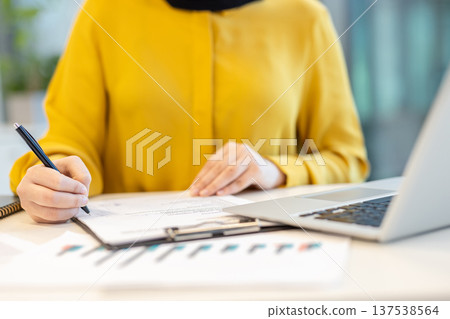 Business woman dressed in yellow top sitting at office desk, holding a pen and signing official papers, with a laptop and financial charts nearby, representing productivity and work 137538564