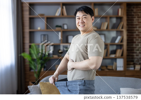 Man celebrating success in weight loss by holding out his baggy oversized jeans, demonstrating the significant transformation and embracing a healthy lifestyle with a cheerful smile at home 137538579