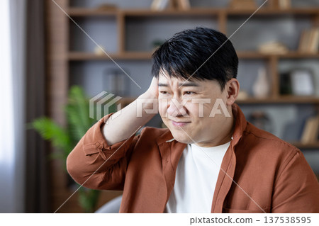 Thoughtful asian man experiencing stress and confusion, holding hand to his head while working in a modern home office setting, contemplating or dealing with a difficult situation Thoughtful asian man experiencing stress and confusion, holding hand to his head while working in a modern home office setting, contemplating or dealing with a difficult situation 137538595