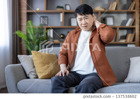 Asian man on gray couch in living room rubbing sore neck with one hand, showing stiffness and discomfort from poor posture or muscle strain while sitting at home Asian man on gray couch in living room rubbing sore neck with one hand, showing stiffness and discomfort from poor posture or muscle strain while sitting at home 137538602