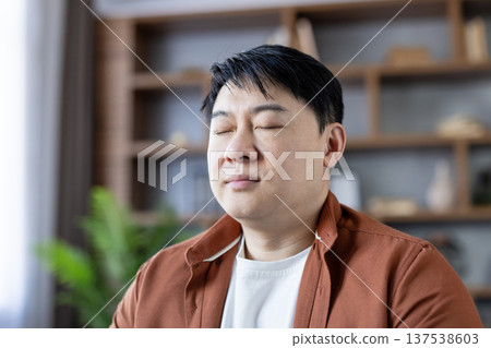 Asian man with closed eyes finding inner balance and feeling relaxed at home, practicing mindfulness, meditation, and mental health for overall well-being 137538603