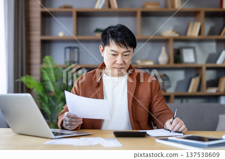 Asian man reviewing documents, writing notes, and calculating expenses, diligently managing personal finances and household budget in his modern home office Asian man reviewing documents, writing notes, and calculating expenses, diligently managing personal finances and household budget in his modern home office 137538609