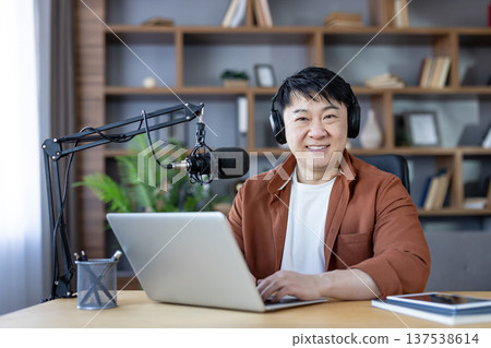 Mature asian man in home studio wearing headphones and smiling while recording an audio podcast with laptop and professional microphone, confident creator broadcasting content Mature asian man in home studio wearing headphones and smiling while recording an audio podcast with laptop and professional microphone, confident creator broadcasting content 137538614