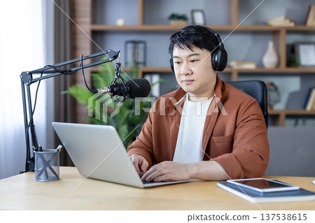 Asian man podcast host wearing headphones and speaking into a studio microphone while typing on a laptop in a modern home office, focused on recording and content creation Asian man podcast host wearing headphones and speaking into a studio microphone while typing on a laptop in a modern home office, focused on recording and content creation 137538615