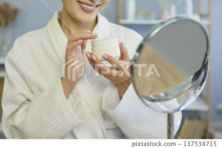 Smiling woman wearing white robe holding jar of cream, trying cosmetics near mirror doing skincare 137538715