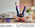 Woman lies on exercise mat and performs core exercise with resistance band around legs 137542508