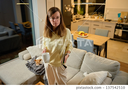 Woman holding coffee mug, tablet and smartphone. Dog resting on sofa 137542510