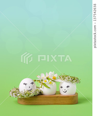 Three white Easter eggs with a cheerful face with white wreath of spring flowers on a gradient green background. Happy Easter greeting card. Three white Easter eggs with a cheerful face with white wreath of spring flowers on a gradient green background. Happy Easter greeting card. 137542638