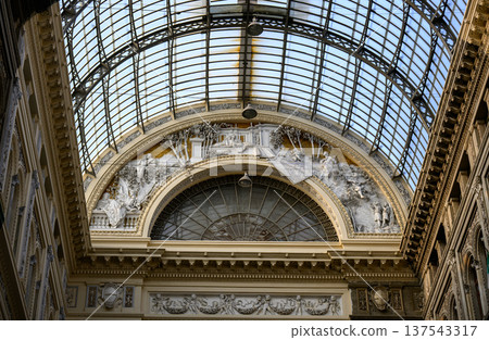 Intricate architecture of Galleria Umberto I in Naples, Italy. The interior features a grand arched glass ceiling supported by metal frames, allowing natural light to illuminate the space. 137543317