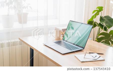 Desk with laptop, notebook, coffee cup, plants near bright window, comfortable remote work environment. Modern home office 137543410