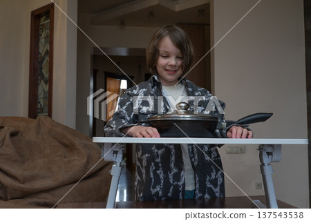 Smiling young boy looking at a covered frying pan on a small table. Child playing chef at home Smiling young boy looking at a covered frying pan on a small table. Child playing chef at home 137543538