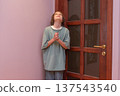 Young boy standing indoors with clasped hands looking up. Child in corner next to a wooden door 137543540
