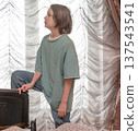 Young boy looking out a window. Caucasian child standing indoors with foot resting on a chair 137543541