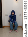 Vertical portrait of a young boy sitting in a corner wearing a blue hooded jacket. Barefoot child 137543544