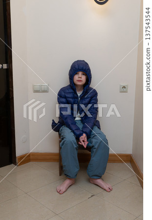 Vertical portrait of a young boy sitting in a corner wearing a blue hooded jacket. Barefoot child Vertical portrait of a young boy sitting in a corner wearing a blue hooded jacket. Barefoot child 137543544