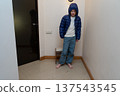 Young barefoot boy in a blue hooded puffer jacket standing indoors. Uncomfortable child grimacing 137543545