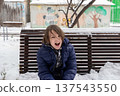 Young boy in blue jacket sitting on a snow-covered bench. Child shouting outdoors in winter 137543550