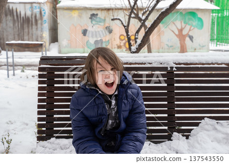 Young boy in blue jacket sitting on a snow-covered bench. Child shouting outdoors in winter Young boy in blue jacket sitting on a snow-covered bench. Child shouting outdoors in winter 137543550