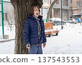 Young boy in blue jacket standing by a tree in the snow. Child shouting outdoors in winter 137543553
