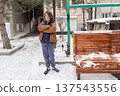 Young boy standing with crossed arms in a snowy urban courtyard. Winter portrait next to a bench 137543556