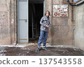 Young boy standing outdoors by an open metal door of a stone building. Winter street portrait 137543558
